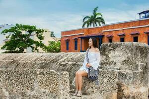 lindo mulher em branco vestir sentado sozinho às a paredes em torno da a colonial cidade do cartagena de índias foto