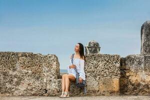 lindo mulher em branco vestir sentado sozinho às a paredes em torno da a colonial cidade do cartagena de índias foto