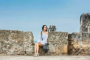 lindo mulher em branco vestir sentado sozinho às a paredes em torno da a colonial cidade do cartagena de índias foto