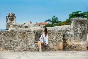 lindo mulher em branco vestir sentado sozinho às a paredes em torno da a colonial cidade do cartagena de índias foto