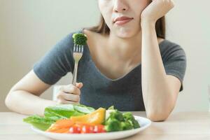 dieta dentro entediado, infeliz lindo ásia jovem mulher, menina em fazer dieta, segurando garfo às brócolis dentro salada placa, não gostar ou cansado com comer fresco legumes. nutrição do limpar, saudável Comida Boa gosto. foto