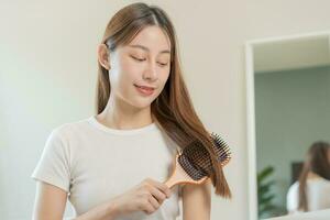 saúde cabelo Cuidado, beleza Maquiagem ásia mulher, menina segurando escova de cabelo e escovar, pentear dela grandes em linha reta cabelo olhando às reflexão dentro espelho dentro manhã rotina depois de salão tratamento, Penteado. foto