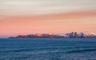 lindo Rosa pôr do sol em a ártico oceano. uma Península com coberto de neve colinas em a horizonte. mar minimalista panorama. foto