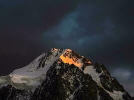 Trevas montanha panorama com ótimo Nevado montanha aceso de alvorecer Sol entre Sombrio nuvens. impressionante alpino cenário com Alto montanha pináculo às pôr do sol ou às nascer do sol. grande geleira em topo dentro laranja claro. foto