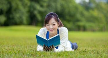 ásia mulher é deitado baixa dentro a Relva gramado dentro a público parque segurando livro dentro dela mão durante verão para lendo e Educação conceito foto