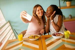 dois jovem mulheres, caucasiano e Preto um, levando selfie com Móvel telefone dentro a cafeteria foto