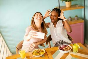 dois jovem mulheres, caucasiano e Preto um, levando selfie com Móvel telefone dentro a cafeteria foto