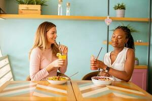 jovem Preto e caucasiano mulher tendo Boa tempo, bebendo fresco sucos e tendo saudável café da manhã dentro a cafeteria foto
