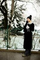 sorridente jovem mulher dentro acolhedor roupas usando Móvel telefone e segurando café copo em inverno dia foto