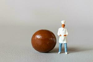 miniatura pessoas chefe de cozinha com chocolate em pé enquanto em pé contra uma cinzento fundo foto