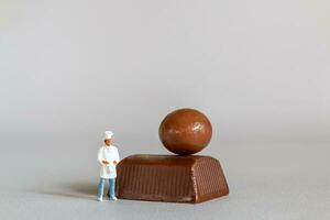 miniatura pessoas chefe de cozinha com chocolate em pé enquanto em pé contra uma cinzento fundo foto