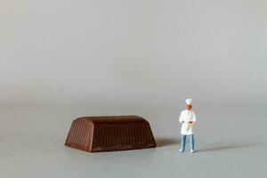 miniatura pessoas chefe de cozinha com chocolate em pé enquanto em pé contra uma cinzento fundo foto