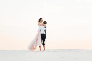 casal jovem um cara de calça preta e uma garota de vestido rosa estão caminhando na areia branca foto