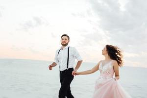 casal jovem um cara de calça preta e uma garota de vestido rosa estão caminhando na areia branca foto