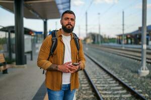 adulto homem é em pé às estrada de ferro estação e esperando para chegada do trem. foto