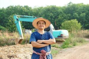 bonito ásia homem agricultor desgasta chapéu, azul camisa, cruzada braços em peito sentir confiante, escavado lagoa de escavadora às agrícola terra para loja água durante seca foto