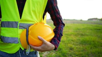 construção trabalhador segurando dele amarelo capacete, Difícil chapéu e construção plano vestindo fluorescente colete em a esvaziar campo. ocupacional segurança e saúde osh foto