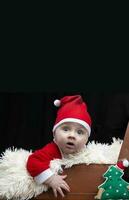 engraçado Natal criança dentro uma santa claus chapéu dentro uma retro mala de viagem em uma Preto fundo. criança dentro Natal. foto