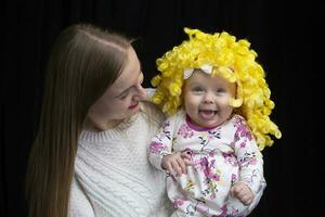 mãe detém dentro dela braços uma pequeno feliz filha dentro uma amarelo peruca. engraçado crianças. foto