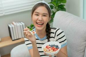 saudável Comida. lindo fêmea menina apreciar comer iogurte, granola, fresco frutas em café da manhã saúde dentro casa. feliz jovem mulher sorrir em manhã Boa emoção. fazer dieta, desintoxicação, dieta, limpar \ limpo comer, vegetariano foto