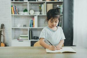 ásia bebê menina vestindo uma azul listrado camisa escrever notas dentro caderno para estude conectados em madeira mesa escrivaninha dentro vivo quarto às lar. Educação Aprendendo conectados a partir de casa conceito. foto