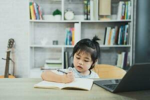 menina asiática vestindo uma camisa listrada azul usa laptop e escreve notas no caderno para estudar on-line na mesa de madeira na sala de estar em casa. educação aprendendo on-line a partir do conceito de casa. foto