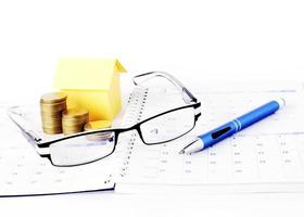 conceito de empréstimos com óculos e caneta azul com moedas de dinheiro e casa de papel amarela na página do calendário foto