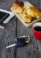 feche a chave do carro e a xícara de café vermelha com pãezinhos no fundo da mesa de madeira foto