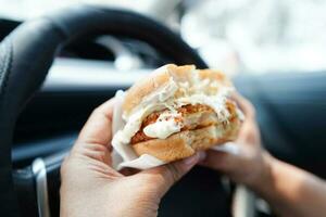 ásia mulher motorista aguarde e comer Hamburger dentro carro, perigoso e risco a acidente. foto