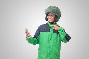 retrato do ásia conectados Táxi motorista vestindo verde Jaqueta e capacete segurando Móvel telefone e apontando para esvaziar espaço com dedo. isolado imagem em branco fundo foto