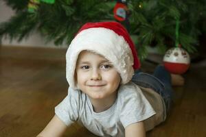 uma foto do uma lindo Garoto dentro uma cinzento camiseta e uma santa claus chapéu às a Natal árvore, olhando para dentro a Câmera. retrato dentro uma brilhante sala. natural, não encenado fotografia.