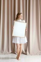 mulher dentro vestir segurando em branco quadro, bege cortina fundo foto