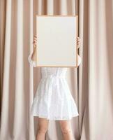mulher dentro vestir segurando em branco quadro, bege cortina fundo foto