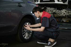 jovem masculino ásia profissional automotivo mecânico trabalhador Verificações pneu pressão de Smartphone inscrição às uma carro garagem, especialista dentro manutenção veículo serviço, e fixação ocupações auto indústria. foto