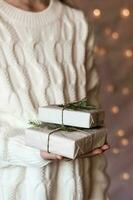 uma menina dentro uma branco tricotado suéter detém presentes dentro papel com uma abeto árvore em uma fundo do luzes. eco amigáveis minimalista Natal. vertical Visão foto