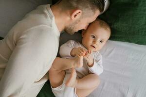 alegre, lindo recém-feito Papai e uma encantador criança de seis meses bebê mentira juntos em verde e branco roupa de baixo. uma jovem pai com uma pequeno criança dentro uma casa retrato. topo Visão foto