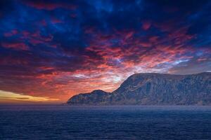 Visão a partir de a mar para a oeste capa dentro Noruega às pôr do sol com Sol raios e pesado nuvens foto