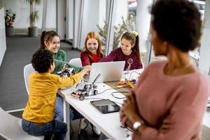 crianças felizes com sua professora de ciências afro-americana com laptop programando brinquedos elétricos e robôs na sala de aula de robótica foto