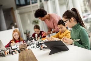 crianças felizes com sua professora de ciências afro-americana com laptop programando brinquedos elétricos e robôs na sala de aula de robótica foto