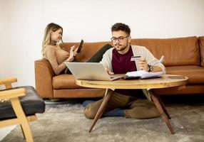 mulher e homem jovem usando laptop para pagamento on-line enquanto estão sentados no sofá em casa