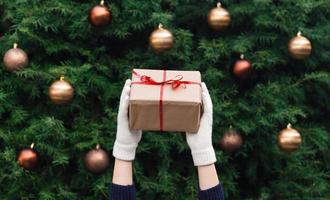 mãos femininas com luvas brancas de malha segurando um presente de Natal embrulhado em papel artesanal foto