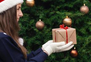 uma jovem com um chapéu de Papai Noel dá um presente feito de papel artesanal com uma fita vermelha foto