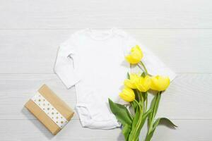 branco brincar recém-nascido camisa. de madeira fundo, amarelo tulipas, feliz aniversário presente caixa. em branco modelo macacão maiô frente topo visualizar. bebê roupas. ramalhete do flores foto