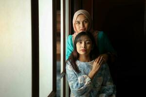 dois jovem ásia malaio muçulmano mulher vestindo lenço de cabeça baju Kurung vestir às casa dentro frente janela humor luz em face Sombrio fundo pacífico conteúdo calma tranquilo pausa cocho maravilha feliz foto