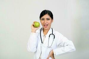 jovem ásia fêmea médico vestindo avental uniforme túnica estetoscópio segurando apontando mostrando comendo saudável verde maçã foto