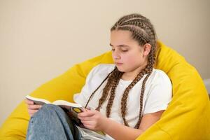 Adolescência menina com tranças lendo livro em amarelo beanbag cadeira foto