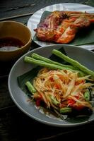 tailandês Comida prato ambos dentro Tailândia e Ásia, mamão salada ou Como nós ligar isto somtum é complementado com grelhado frango e pegajoso arroz com fresco frituras. servido em a Preto de madeira mesa. foto