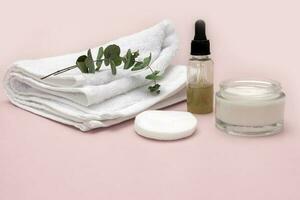 Cosmético acessórios em uma pálido Rosa fundo, uma toalha, sérum, face creme, algodão almofadas, uma raminho do vegetação foto