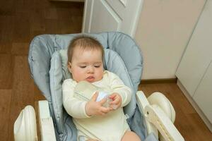 bebê menina em uma alimentando cadeira parece às uma garrafa com uma mistura com interesse foto
