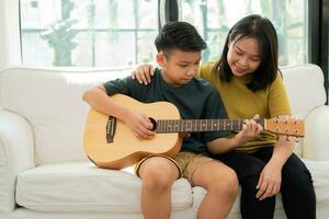 mãe asiática abraça filho, menino asiático tocando violão e mãe abraçada no sofá e se sente apreciada e encorajada. conceito de uma família feliz, aprendizado e estilo de vida divertido, ama os laços familiares foto